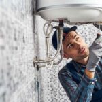 A plumber inspects a heating system in a tiled bathroom, using a flashlight and wrench, wearing gloves and a plaid shirt.