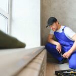 A worker in a blue uniform and cap installs or repairs a gas line in a room with gray walls and a large window.