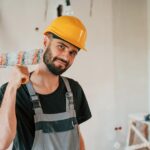 A construction worker wearing a yellow hard hat playfully poses with a paint roller.